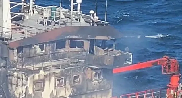 O navă cu urme de arsură pe suprastructură navighează pe un mar agitat sub un cer înnorat.