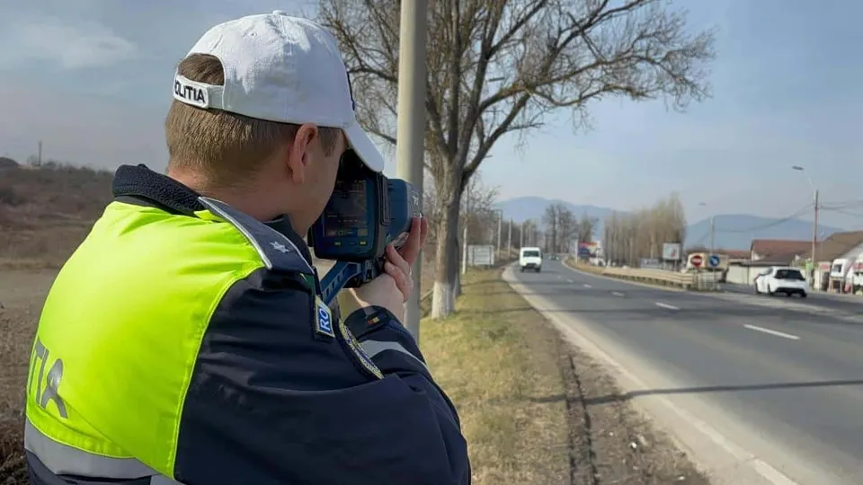 Un agent de poliție cu șapcă albă și vestă reflectorizantă folosește un aparat de măsurat viteza pe marginea unui drum asfaltat cu mașini în depărtare.
