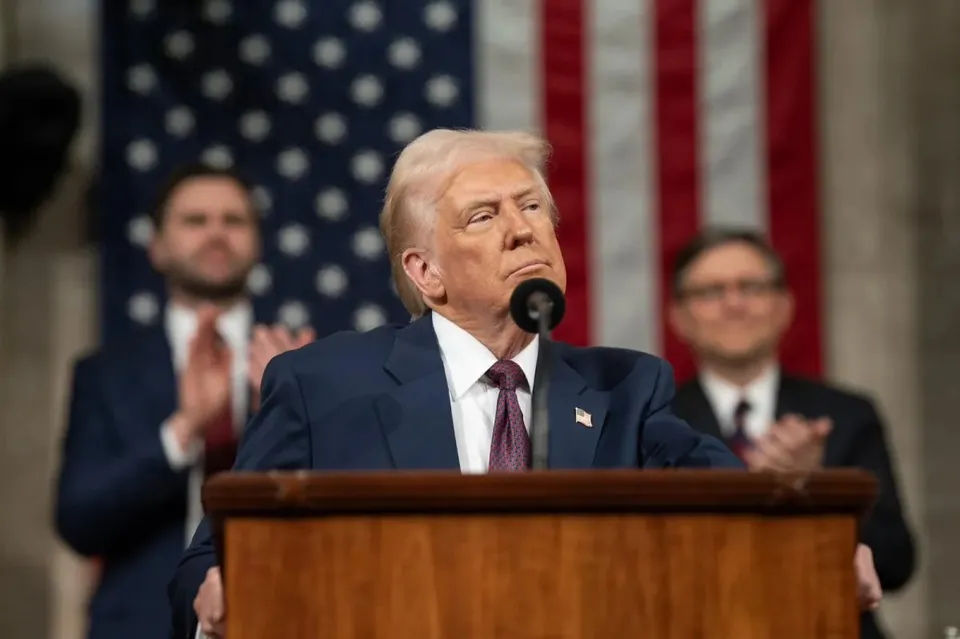Un bărbat cu părul blond, îmbrăcat în costum închis și cravată purpurie, stă în spatele unui podium de lemn în fața unui steag american.