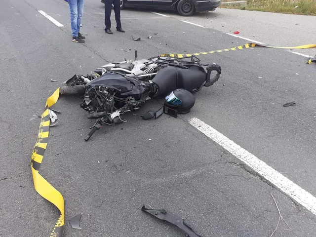 O motocicletă neagră este întinsă pe asfaltul unei străzi lângă un cască de protecție și o bandă de delimitare galbenă și neagră.