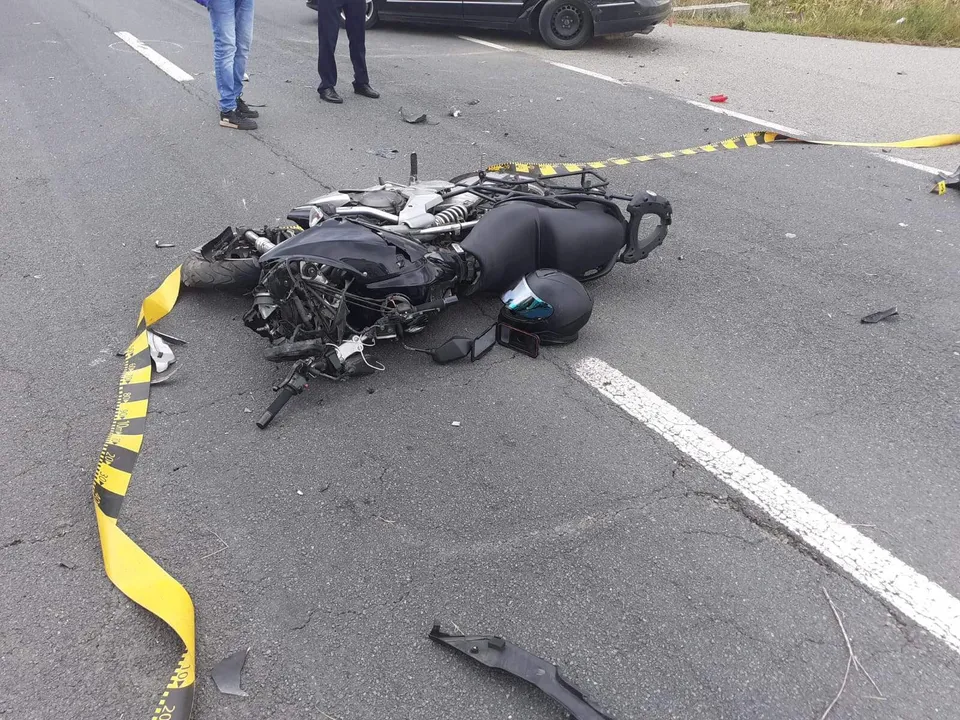 O motocicletă neagră este întinsă pe asfaltul unei străzi lângă un cască de protecție și o bandă de delimitare galbenă și neagră.