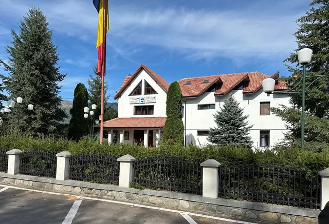 O clădire albă cu acoperiș de țiglă roșie este situată în spatele unui gard negru și a unor copaci verzi sub un cer albastru.