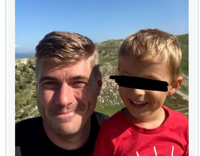 Un bărbat cu părul scurt și un copil cu tricou roșu stau unul lângă altul în fața unui peisaj muntos sub cerul senin.