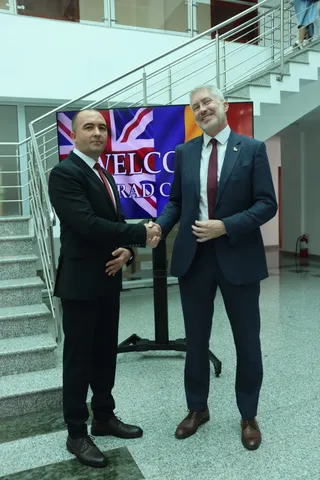 Doi bărbați în costume îmbrăcați stau în picioare și se strâng în mână în fața unui panou colorat într-un spațiu interior cu scări metalice.