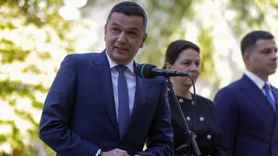 Un bărbat într-un costum albastru și cravată stă în fața unui microfon, fiind însoțit de alte două persoane în plan secund.