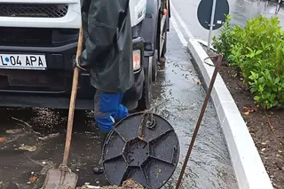 Un bărbat într-un halat negru și pantaloni albaștri ține o lopată lângă un vehicul alb și un capac de canalizare pe o stradă umedă.