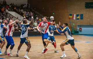 Cinci jucători de handbal în echipamente colorate participă la un meci pe un teren de interior în fața unor tribune cu spectatori.