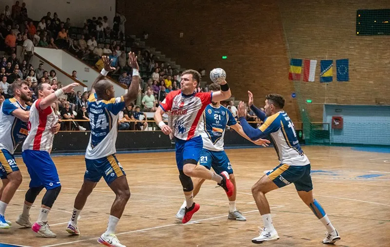 Cinci jucători de handbal în echipamente colorate participă la un meci pe un teren de interior în fața unor tribune cu spectatori.