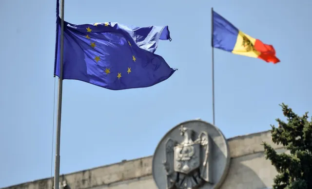Două steaguri, unul albastru cu stele galbene și unul tricolor, flutură pe catarge sub un cer senin deasupra unei clădiri cu un emblema sculptată.