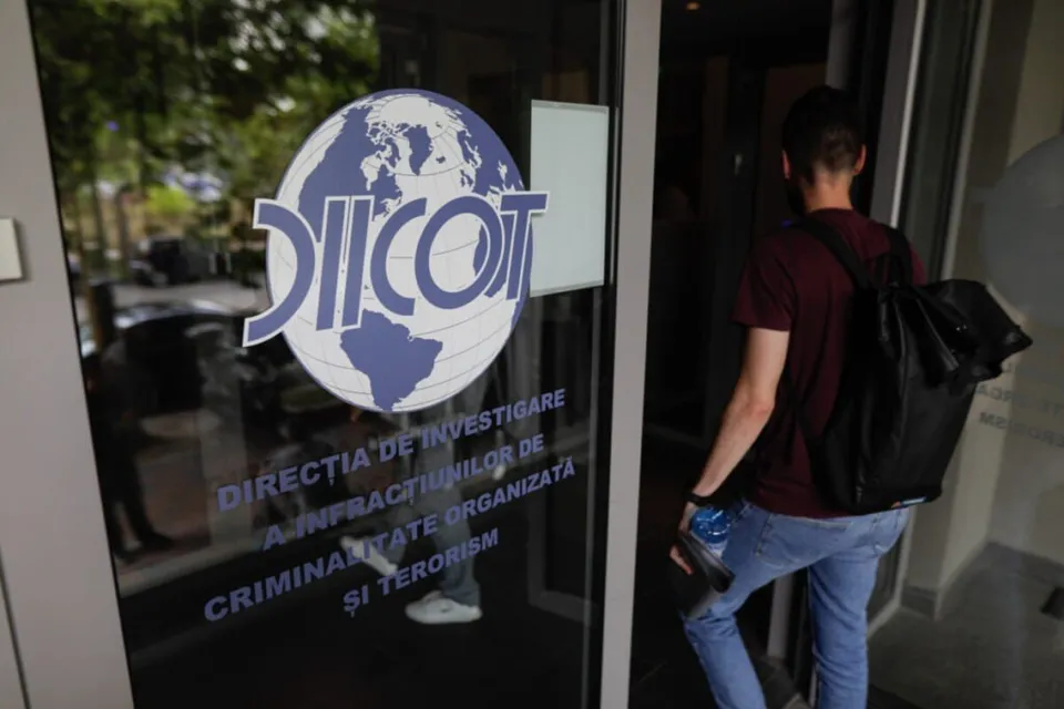 Un bărbat cu rucsac negru și tricou bordeaux pășește printr-o ușă de sticlă pe care este aplicat un logo albastru cu textul DIICOT.