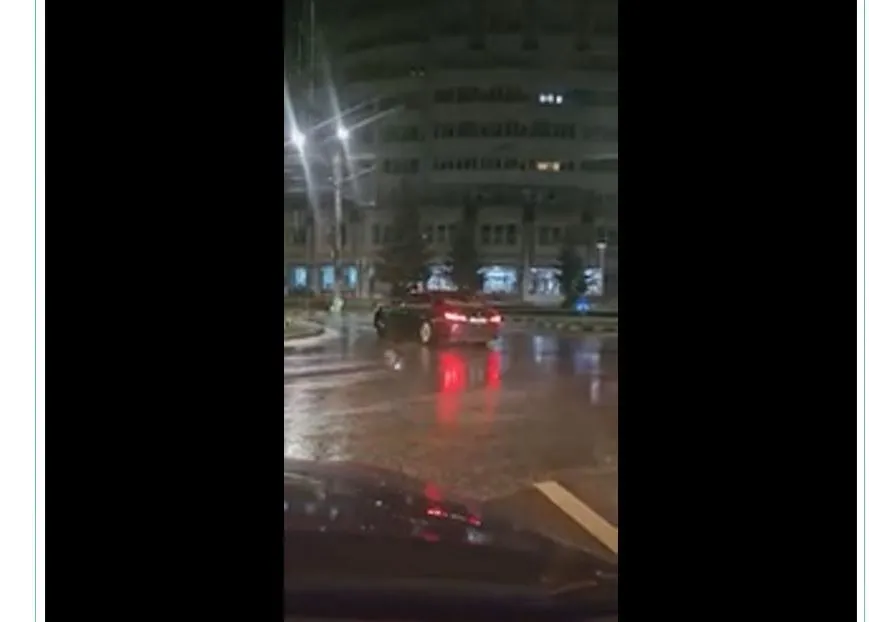 O mașină roșie circulă pe un drum umed noaptea în fața unei clădiri mari cu ferestre iluminate.