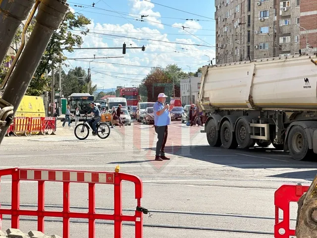 Un bărbat stă pe o stradă lângă un camion mare, un biciclist trece pe lângă bariere roșii și albe, iar vehicule sunt parcate în fundal.
