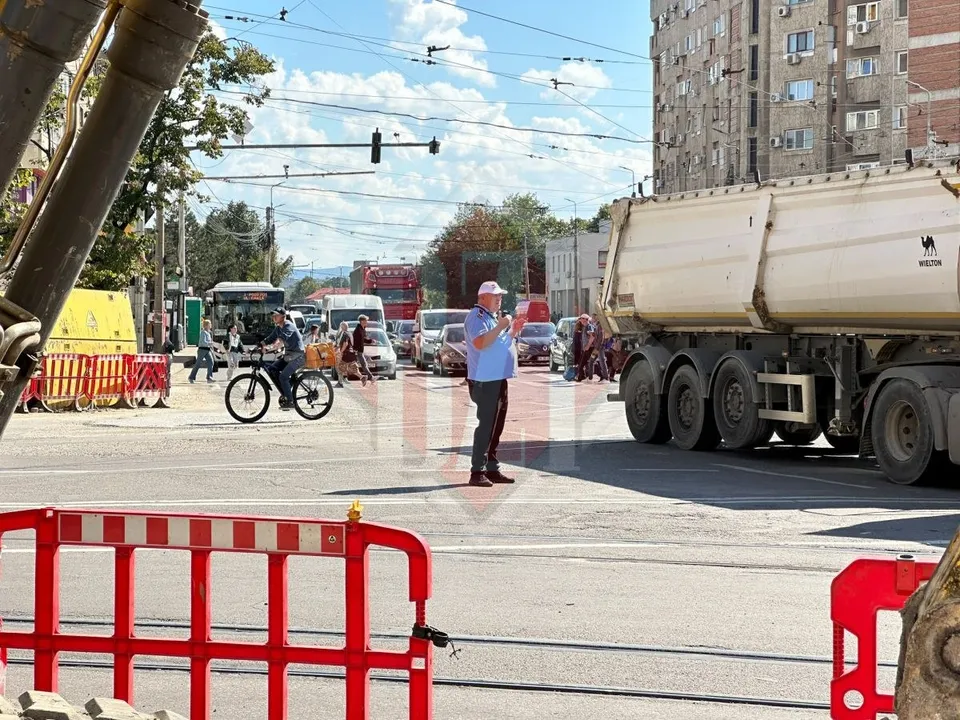 Un bărbat stă pe o stradă lângă un camion mare, un biciclist trece pe lângă bariere roșii și albe, iar vehicule sunt parcate în fundal.