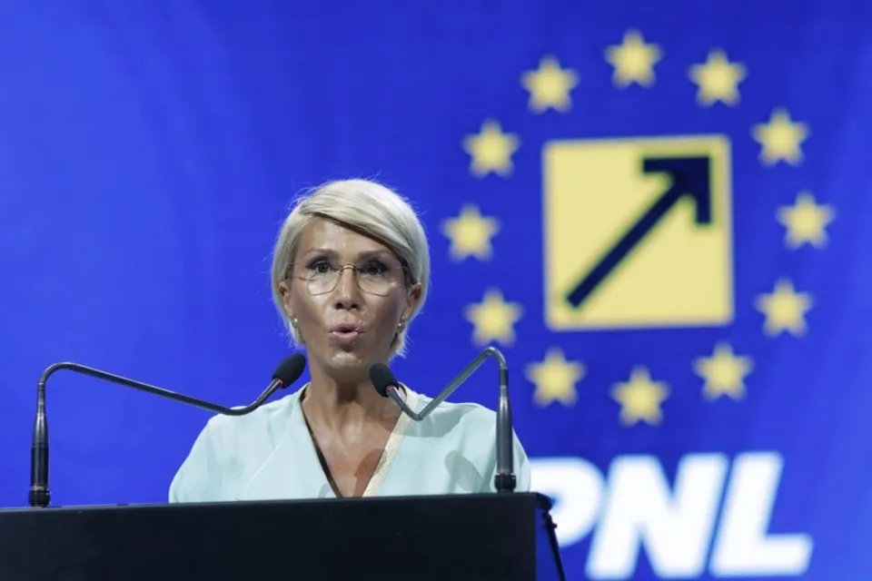 O femeie cu părul scurt și deschis la culoare stă în spatele unui podium cu microfoane, având un fundal albastru cu stele și un logo galben.