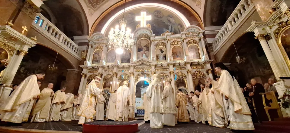 Oamenii în haine albe stau în rând în fața unui iconostas bogat decorat într-o biserică cu un candelabru suspendat.