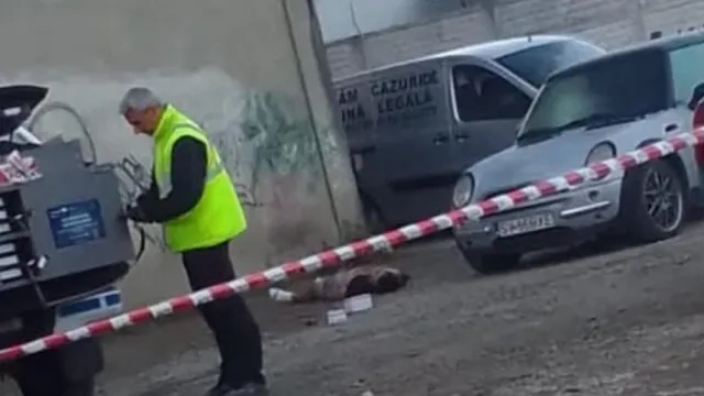 Un bărbat cu vestă reflectorizantă verde stă lângă un echipament tehnic, în timp ce o bandă de delimitare albă și roșie traversează scena.
