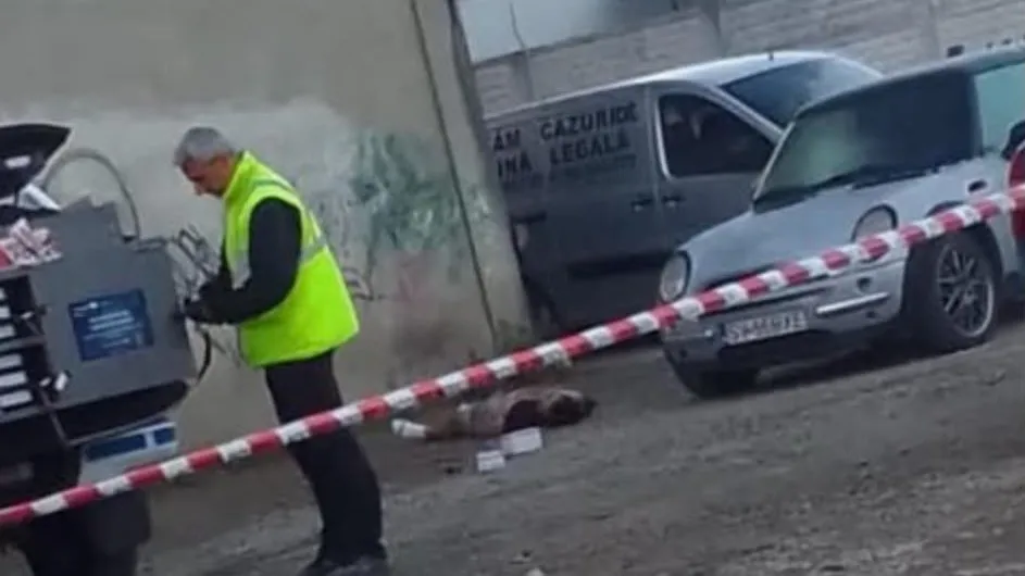 Un bărbat cu vestă reflectorizantă verde stă lângă un echipament tehnic, în timp ce o bandă de delimitare albă și roșie traversează scena.