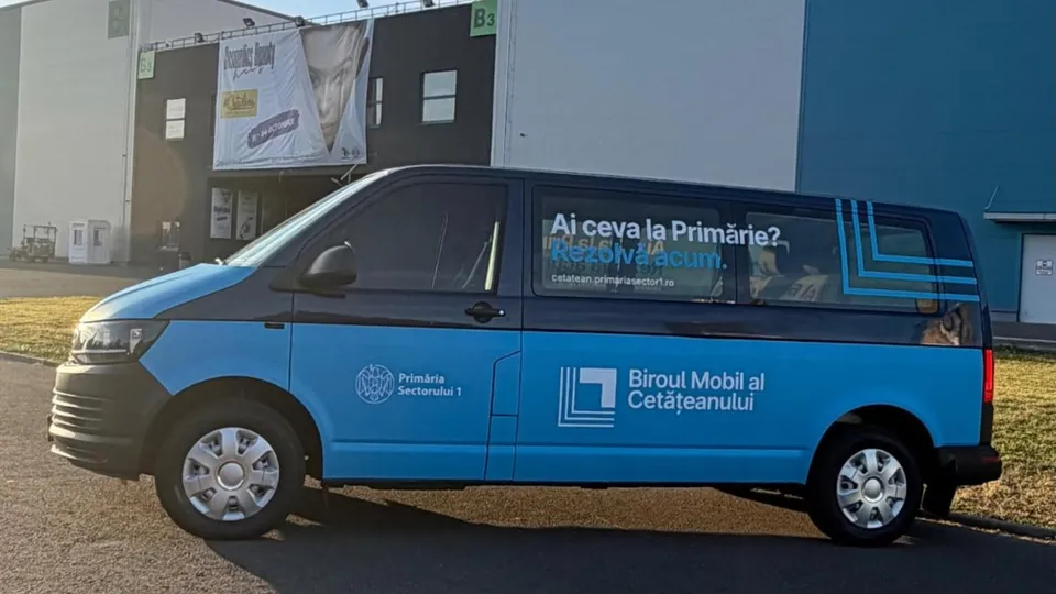 Un van albastru și negru cu inscripții pe lateral este parcat în fața unei clădiri cu un panou publicitar suspendat.