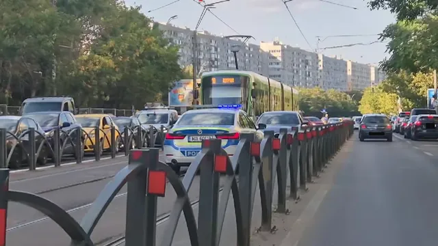Un tramvai verde și galben circulă pe șine, în timp ce mai multe autoturisme sunt parcate sau circulă pe o stradă cu clădiri înalte în fundal.