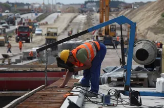 Un bărbat cu cască galbenă și vestă reflectorizantă orange se apleacă pe o suprafață de lemn într-un șantier cu utilaje de construcții.