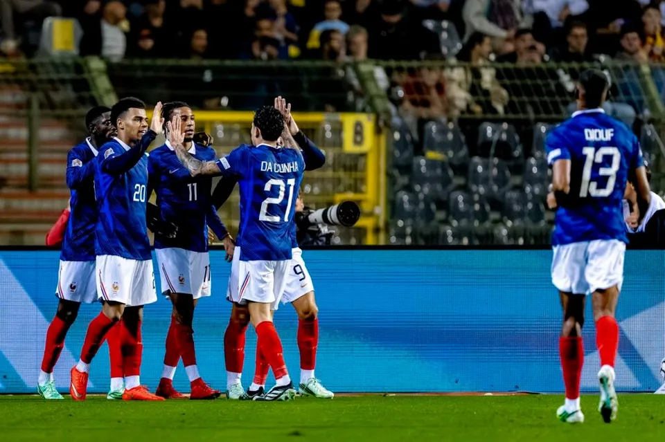 Cinci jucători de fotbal în echipamente albastre și albe sărbătoresc pe un teren de fotbal în fața unor tribune cu spectatori.