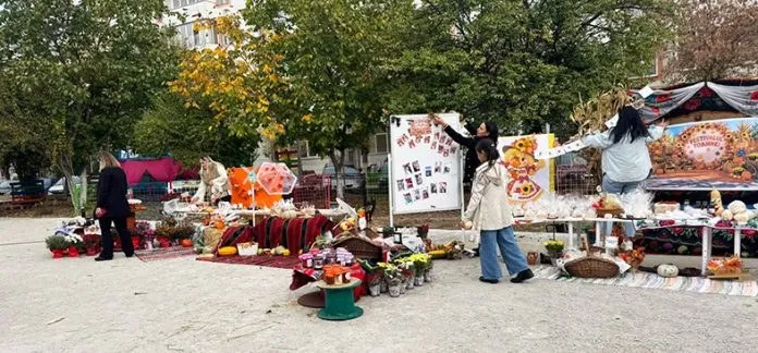 Câțiva oameni stau lângă mese cu diverse produse și decorații de toamnă într-o curte cu copaci verzi și galbeni.