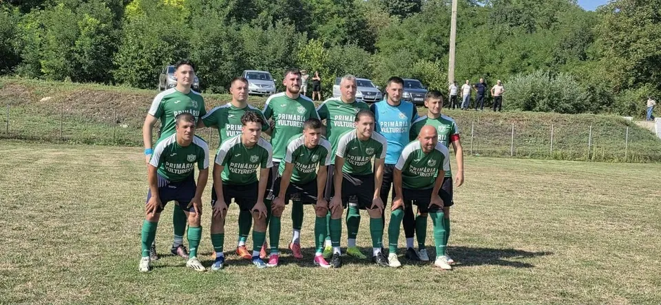 Un grup de unsprezece bărbați îmbrăcați în echipamente sportive verzi și negre pozează pe un teren de fotbal cu copaci în fundal.