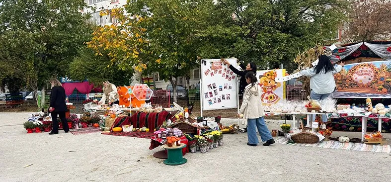 Copii și adulți stau lângă mese cu diverse produse și decorații colorate într-o curte cu copaci verzi și galbeni.