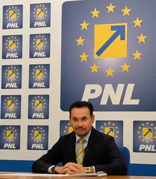 Un bărbat cu părul scurt și costum închis, purtând o cravată galbenă, stă la o masă în fața unui panou albastru cu logo-uri PNL.