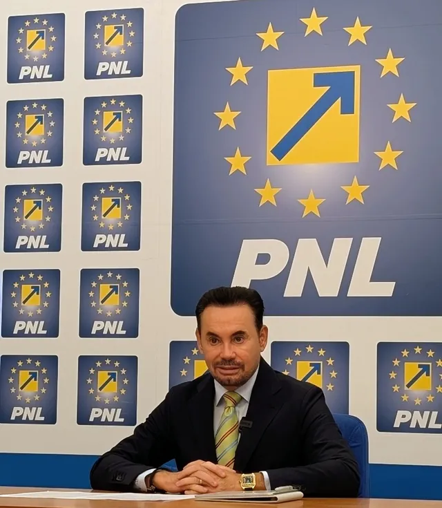 Un bărbat cu părul scurt și costum închis, purtând o cravată galbenă, stă la o masă în fața unui panou albastru cu logo-uri PNL.