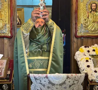 Un bărbat cu barbă poartă haine liturgice verzi și ține un obiect metalic decorat cu pietre colorate în mâinile ridicate.