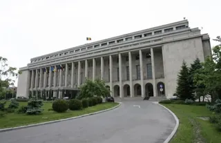 O clădire înaltă de beton cu coloane lungi este înconjurată de vegetație și un drum asfaltat curbat în prim-plan.