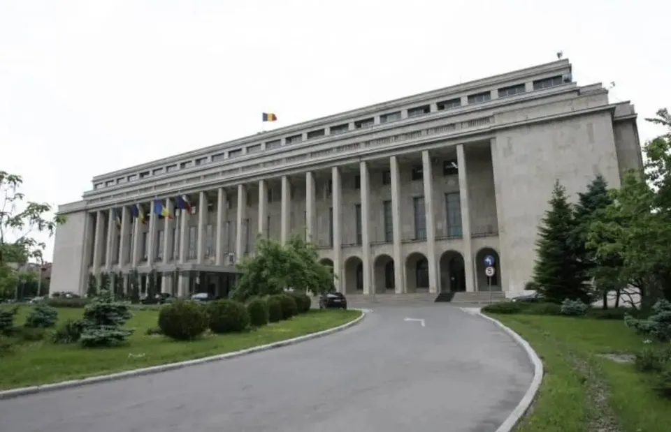O clădire înaltă de beton cu coloane lungi este înconjurată de vegetație și un drum asfaltat curbat în prim-plan.