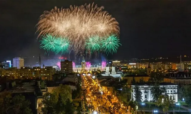 Explozii de artificii colorate de nuanțe aurii și verzi deasupra unei zone urbane iluminate noaptea.