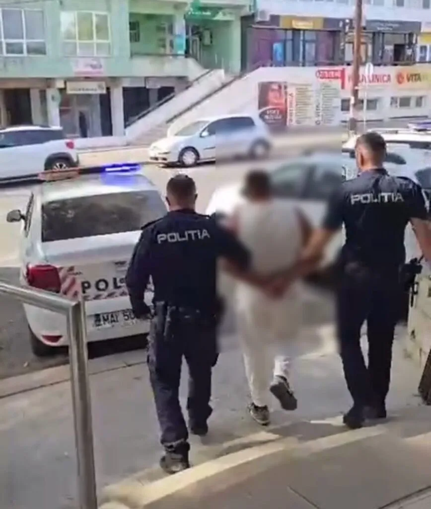 Unii polițiști în uniformă neagră conduc un bărbat cu haine albe pe un trotuar lângă un autoturism alb.