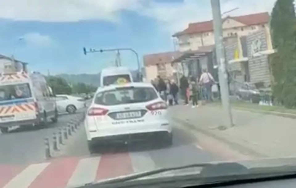 Un autoturism alb este parcat pe marginea drumului lângă un vehicul de serviciu alb și o mulțime de oameni pe trotuar.