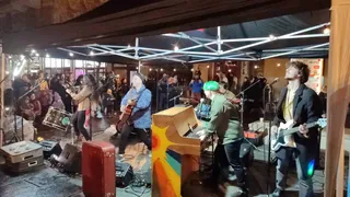 O trupă cântă pe o scenă acoperită, formată din chitarist, basist, pianist și un cântăreț, în fața unui public adunat într-un spațiu interior.