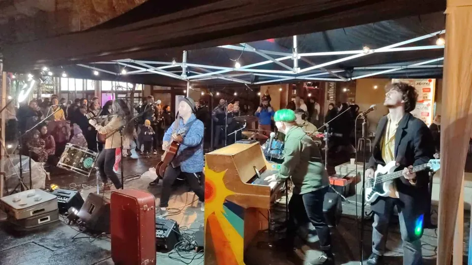 O trupă cântă pe o scenă acoperită, formată din chitarist, basist, pianist și un cântăreț, în fața unui public adunat într-un spațiu interior.