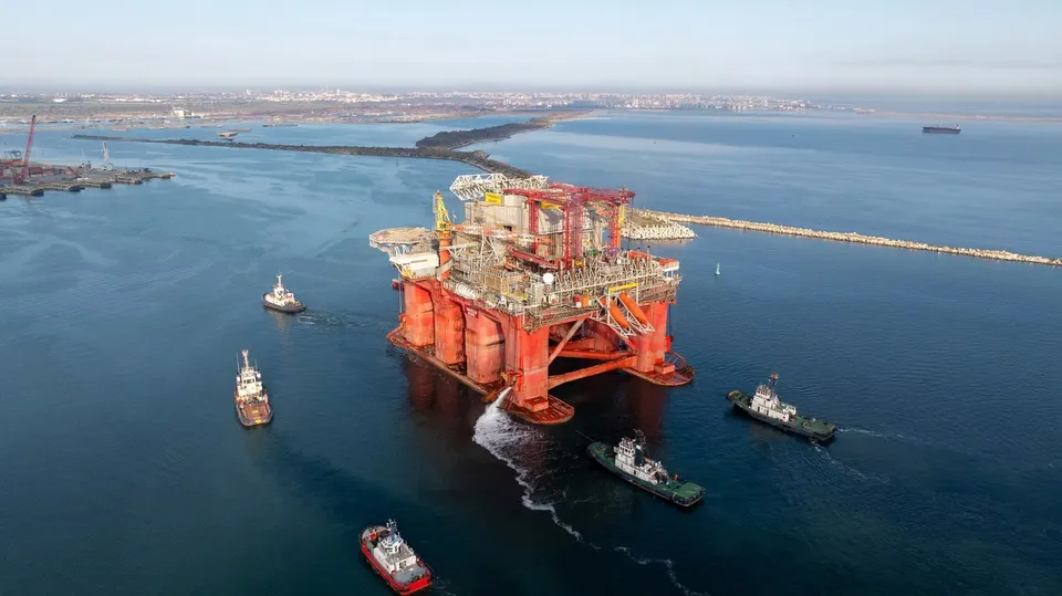 O platformă petrolieră de culoare portocalie este înconjurată de patru nave mici pe o suprafață de apă de culoare albastră sub un cer senin.