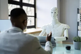Un bărbat stă la o masă și privește un robot alb de dimensiuni medii aflat în fața unui laptop.