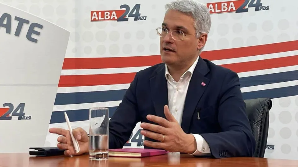 Un bărbat cu ochelari și părul cărunt, purtând sacou închis și cămașă albă, stă la o masă lângă un pahar cu apă și un obiect subțire alb.