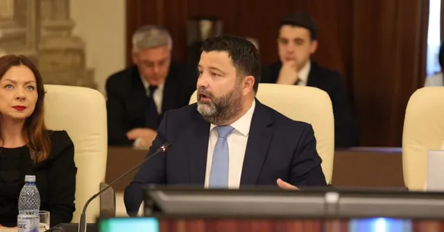 Un bărbat cu barbă, purtând costum închis și cravată deschisă, stă la o masă în fața unui microfon într-o încăpere cu scaune albe.
