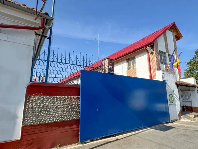 O clădire albă cu acoperiș roșu este delimitată de un gard metalic și un zid cu o porțiță albă și un panou albastru mare.