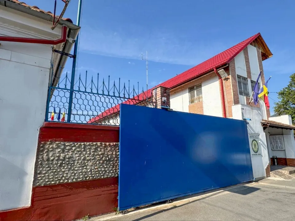 O clădire albă cu acoperiș roșu este delimitată de un gard metalic și un zid cu o porțiță albă și un panou albastru mare.