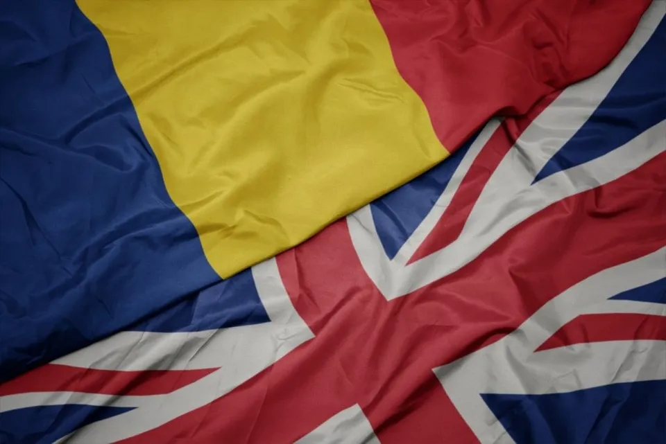 Două steaguri, unul cu dungi verticale albastru, galben și roșu și unul cu modelul Union Jack, sunt întrepătrunse și prezintă pliuri de material textil.