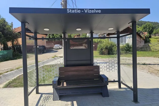 O stație de autobuz cu acoperiș plat, două panouri laterale transparente și o bancă de lemn situată pe un trotuar pavat.