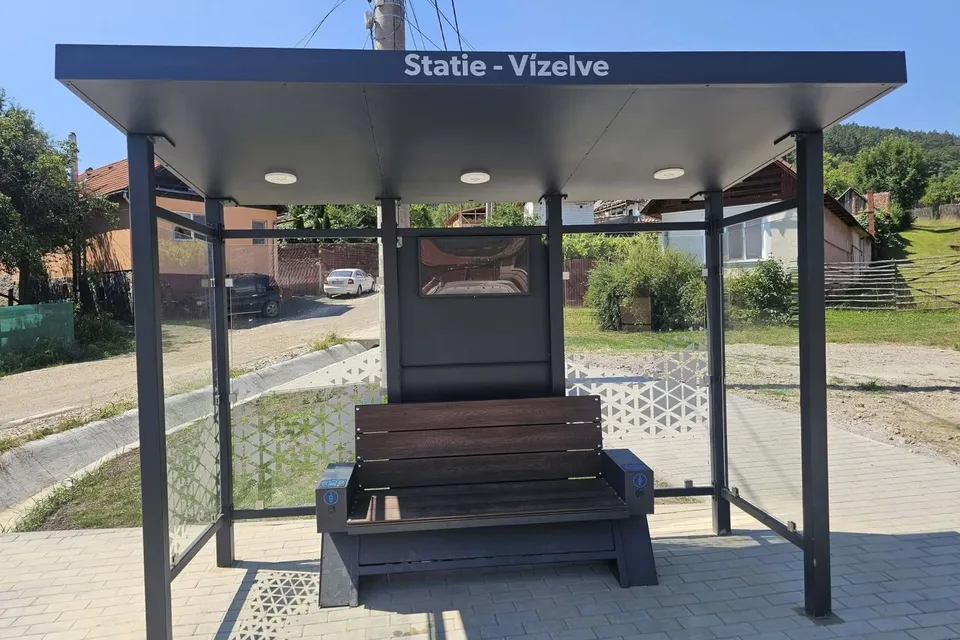 O stație de autobuz cu acoperiș plat, două panouri laterale transparente și o bancă de lemn situată pe un trotuar pavat.