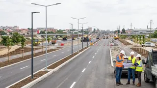 O imagine cu o șoseauă nouă, cu linii proaspete, stâlpi de iluminat și trei persoane în echipament de protecție care discută lângă un autovehicul.