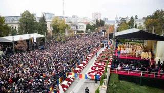 O mulțime mare de oameni este adunată într-o zonă deschisă lângă o platformă ridicată unde stau persoane îmbrăcate în haine albe.