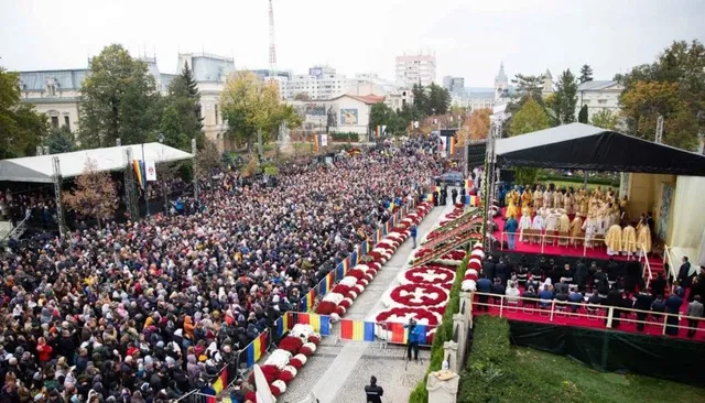 O mulțime mare de oameni este adunată într-o piață publică lângă o scenă cu persoane îmbrăcate în haine albe și roșii.